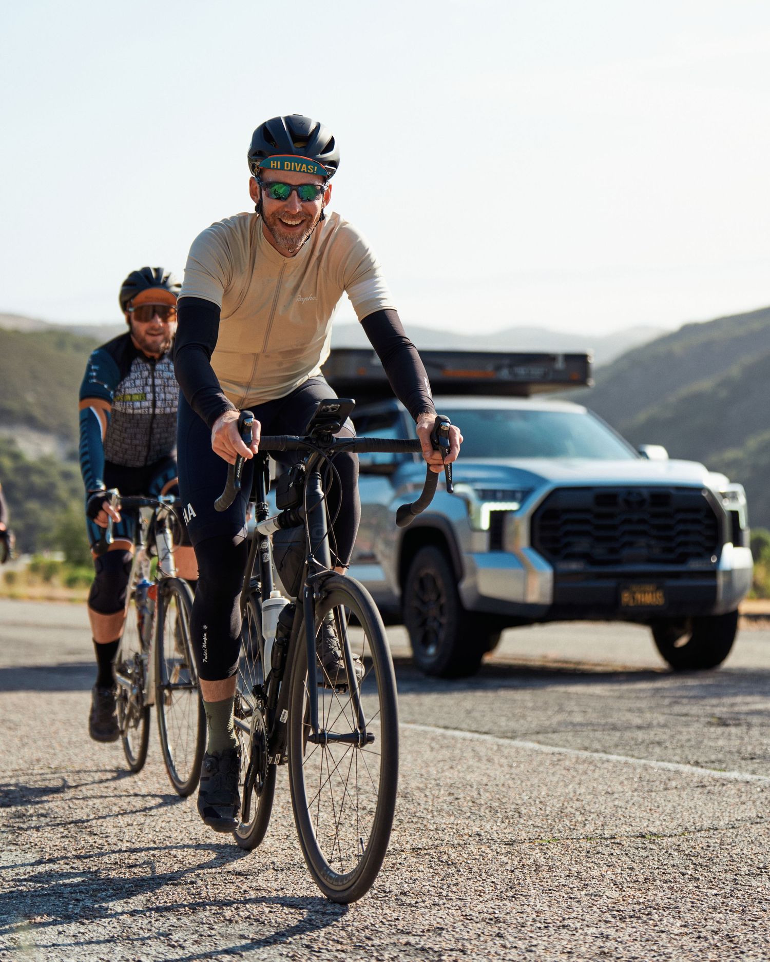 Road cyclists ride past a parked pickup truck on a mountain road wearing cycling apparel and protective gear.