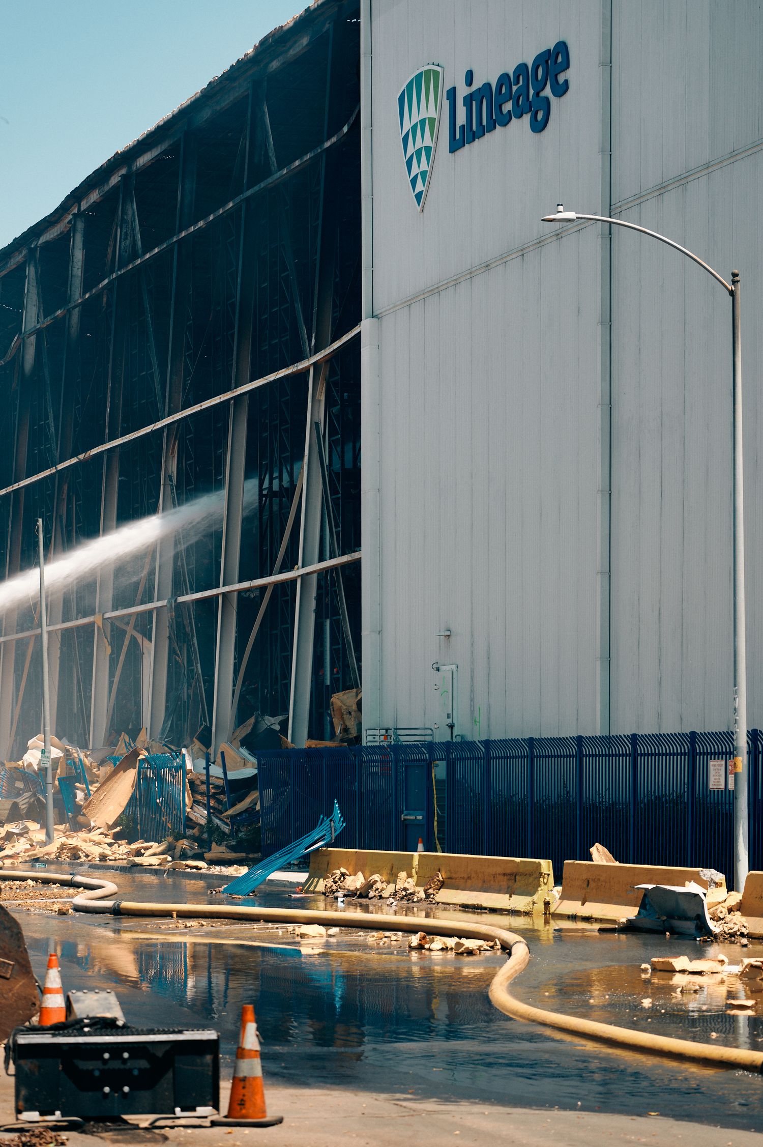 Lineage cold storage warehouse fire with structural damage, fire hoses spraying water, debris scattered, and traffic cones visible.