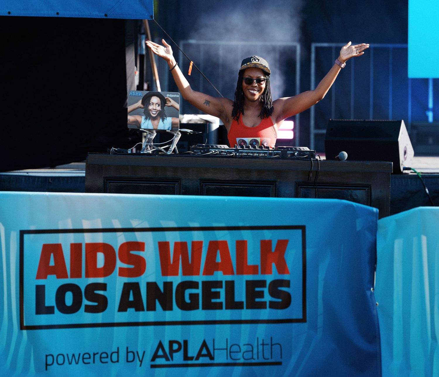 A DJ performs behind turntables at the AIDS Walk Los Angeles event with a blue banner visible.