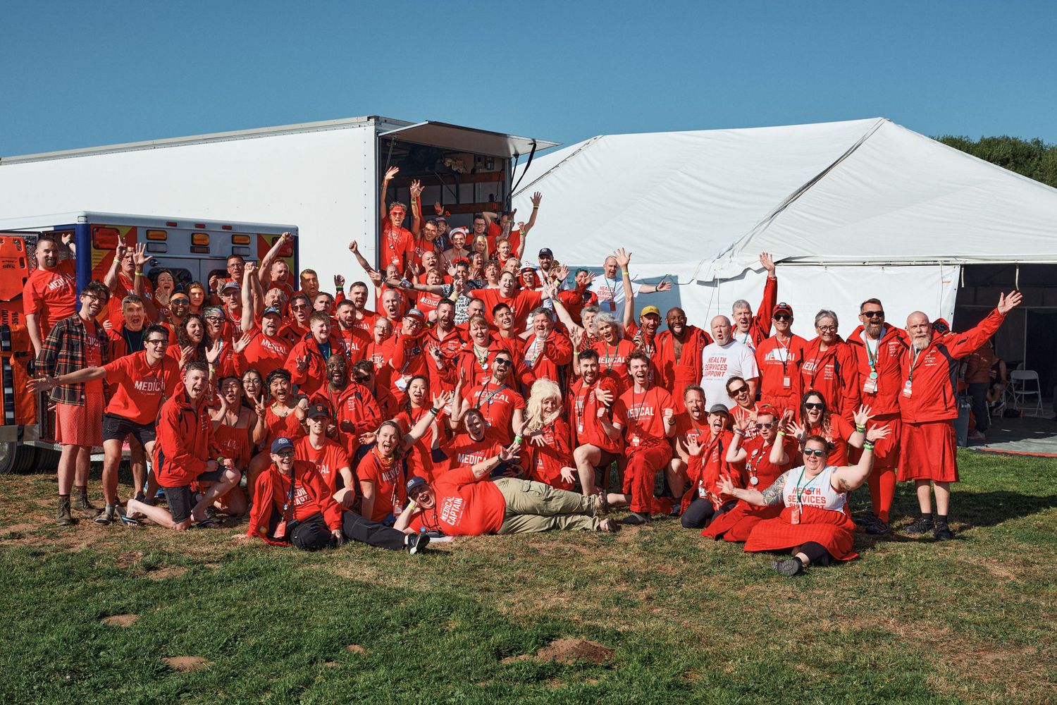 Large crowd dressed in bright red clothing sitting on grass near white tents at an outdoor gathering.