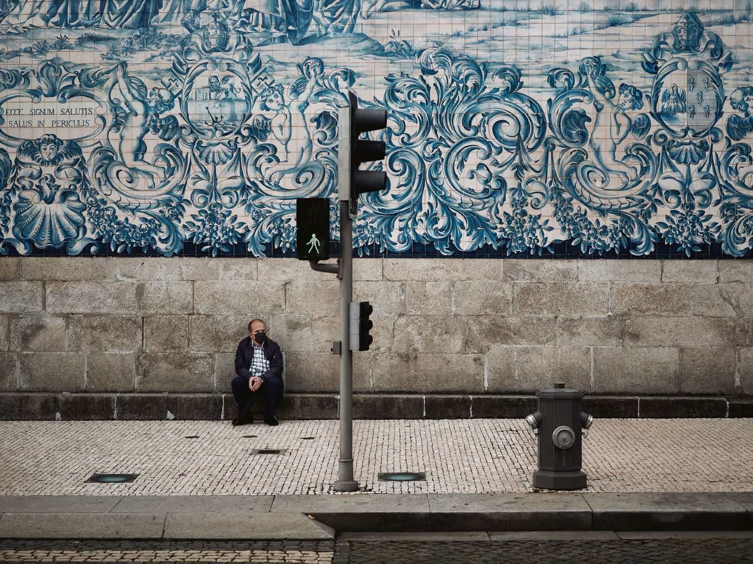 A person sits against a wall decorated with intricate blue and white tile patterns in an urban setting.