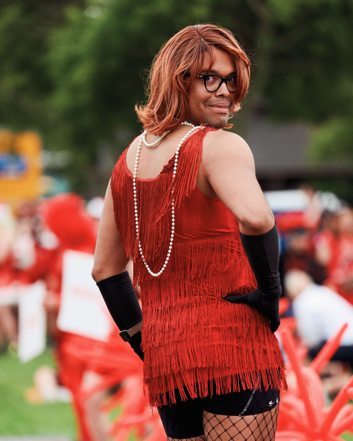Person in red flapper dress with black gloves, pearl necklace and glasses poses playfully at outdoor event.