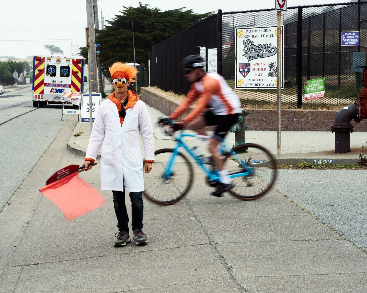 Person in white lab coat and orange mask directs traffic as cyclist rides past on urban street.