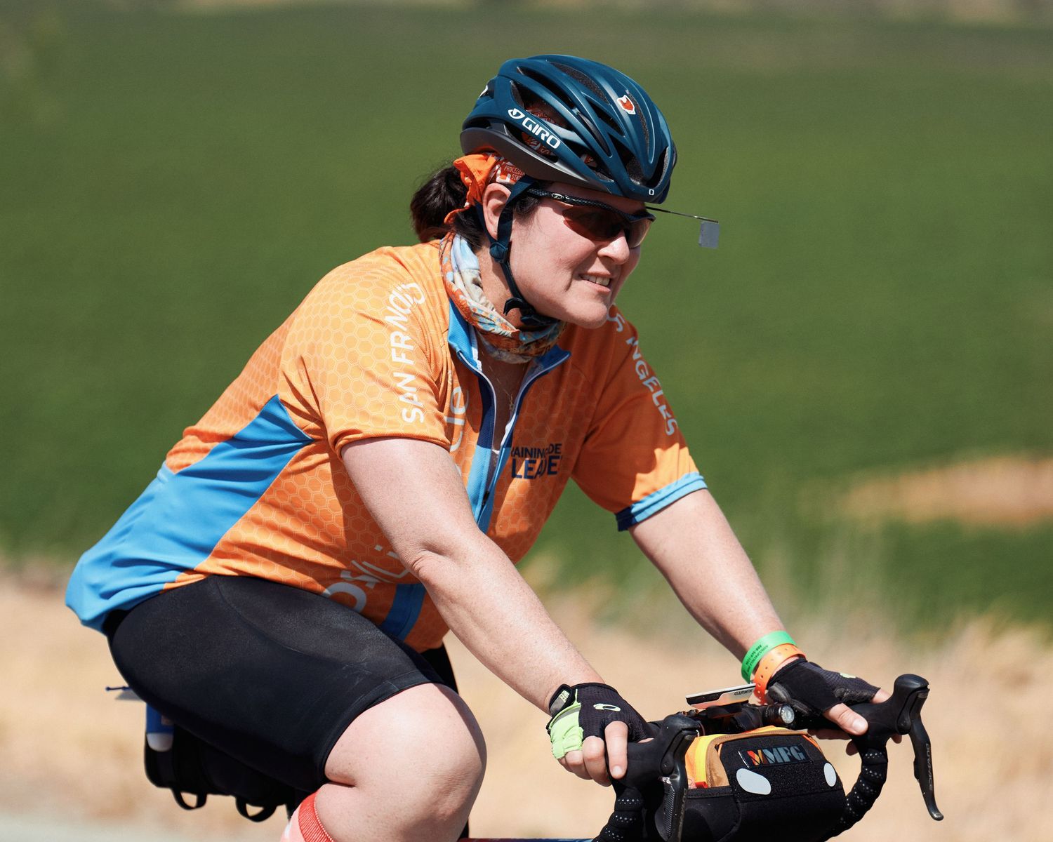 Cyclist in orange and blue jersey pedals along green hillside with handlebar bag.