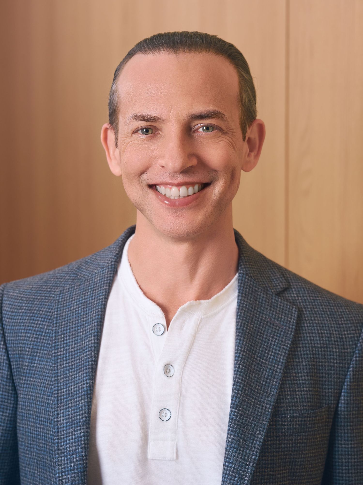 Smiling man in blue blazer and white henley shirt poses for professional headshot against wooden background.
