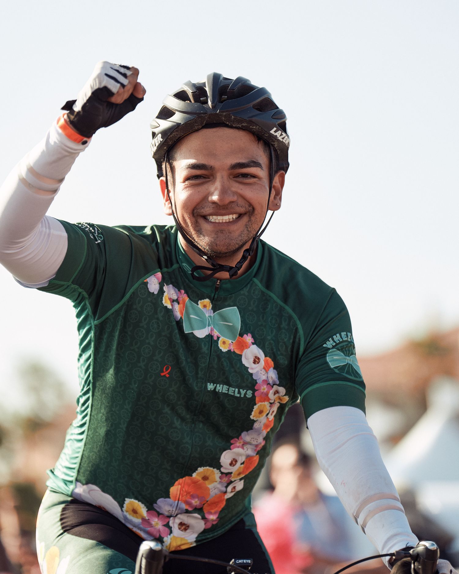A cyclist in a green jersey with floral pattern and cycling helmet raises an arm in celebration during an outdoor event.
