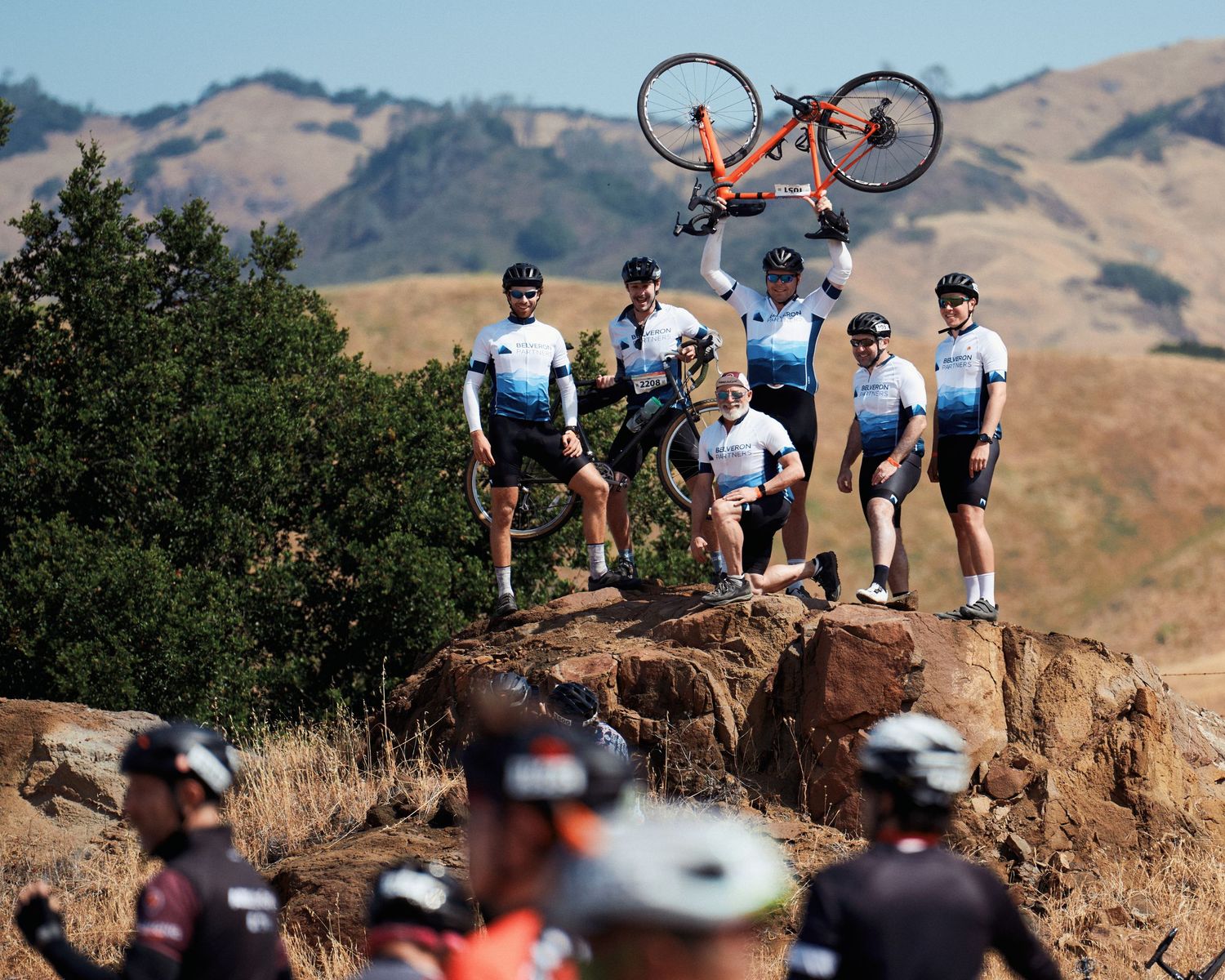 Cyclists gather on a rocky outcrop as one person holds a bicycle overhead against a mountainous backdrop.