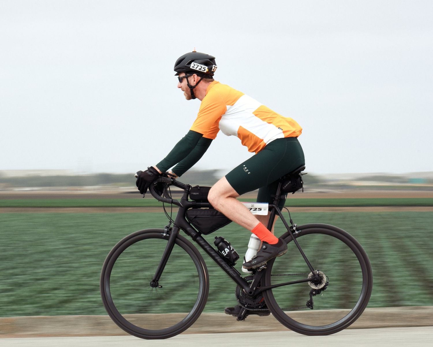 Cyclist wearing bright orange jersey and dark shorts rides black road bike on rural path.