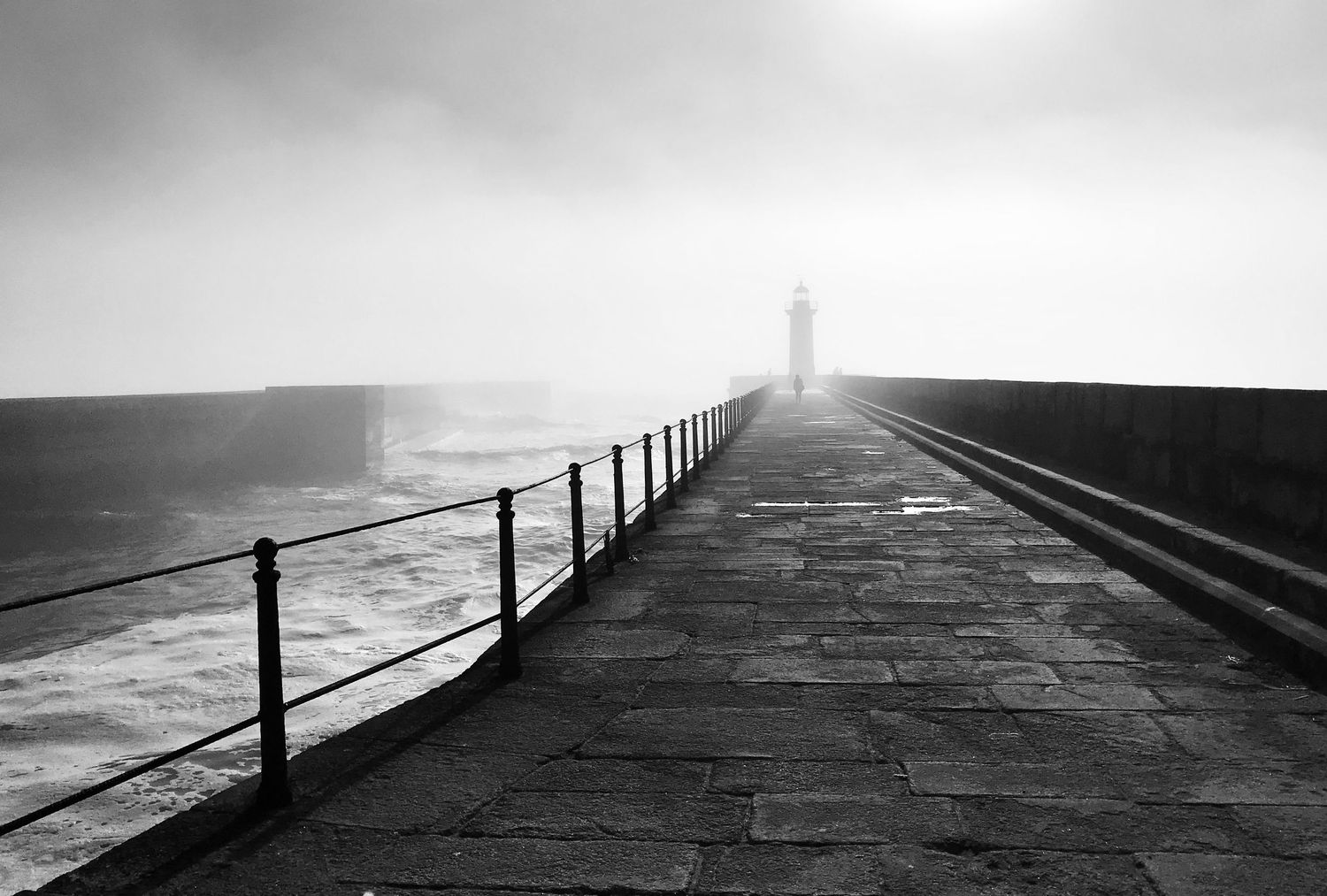 A misty stone pier with metal railings extends into the foggy ocean creating a moody black and white seascape.
