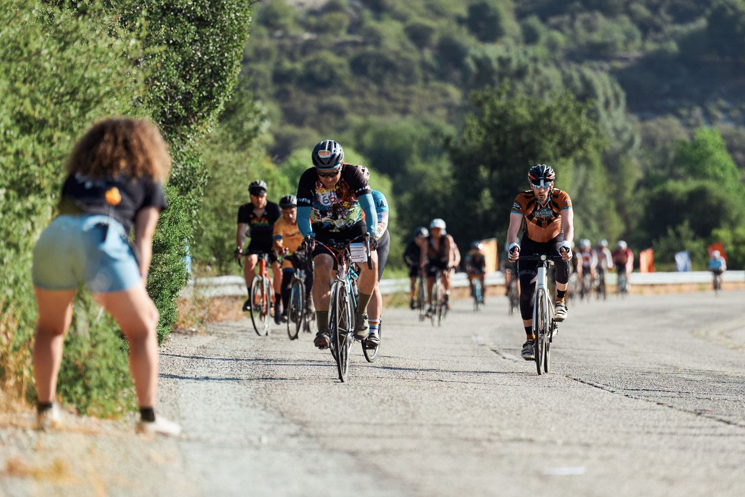A group of cyclists rides along a scenic road while someone photographs them from the side of the path.
