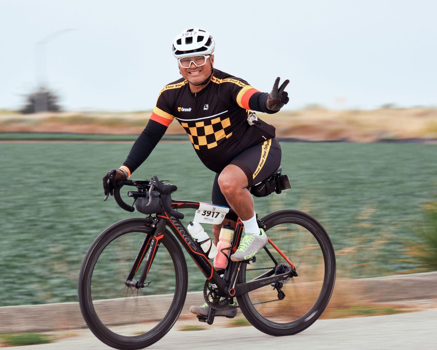 Cyclist in black and orange jersey gesturing peace sign while riding on country road.