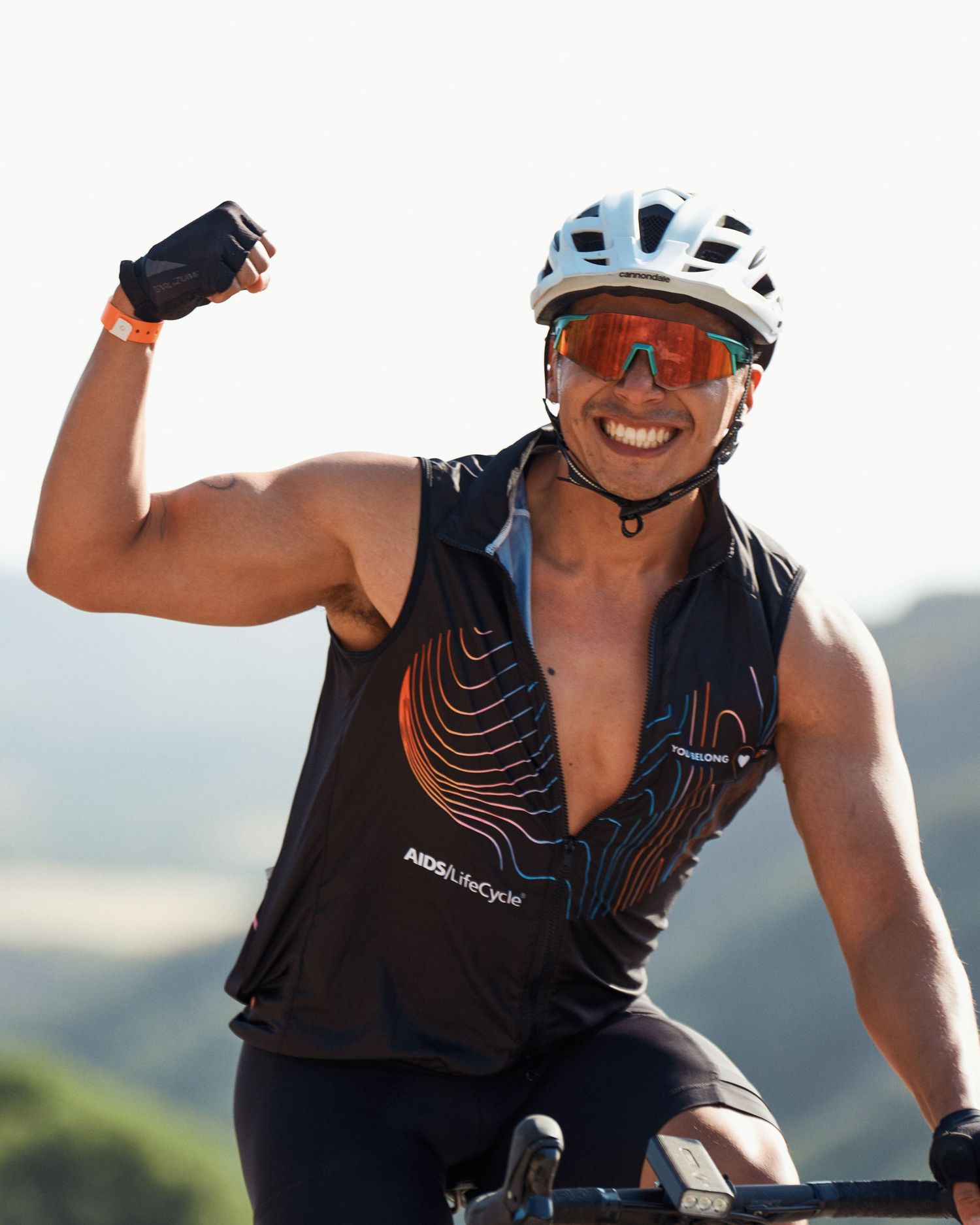 Athletic cyclist in a black jersey celebrates during outdoor ride on sunny mountain trail while wearing sunglasses and helmet.