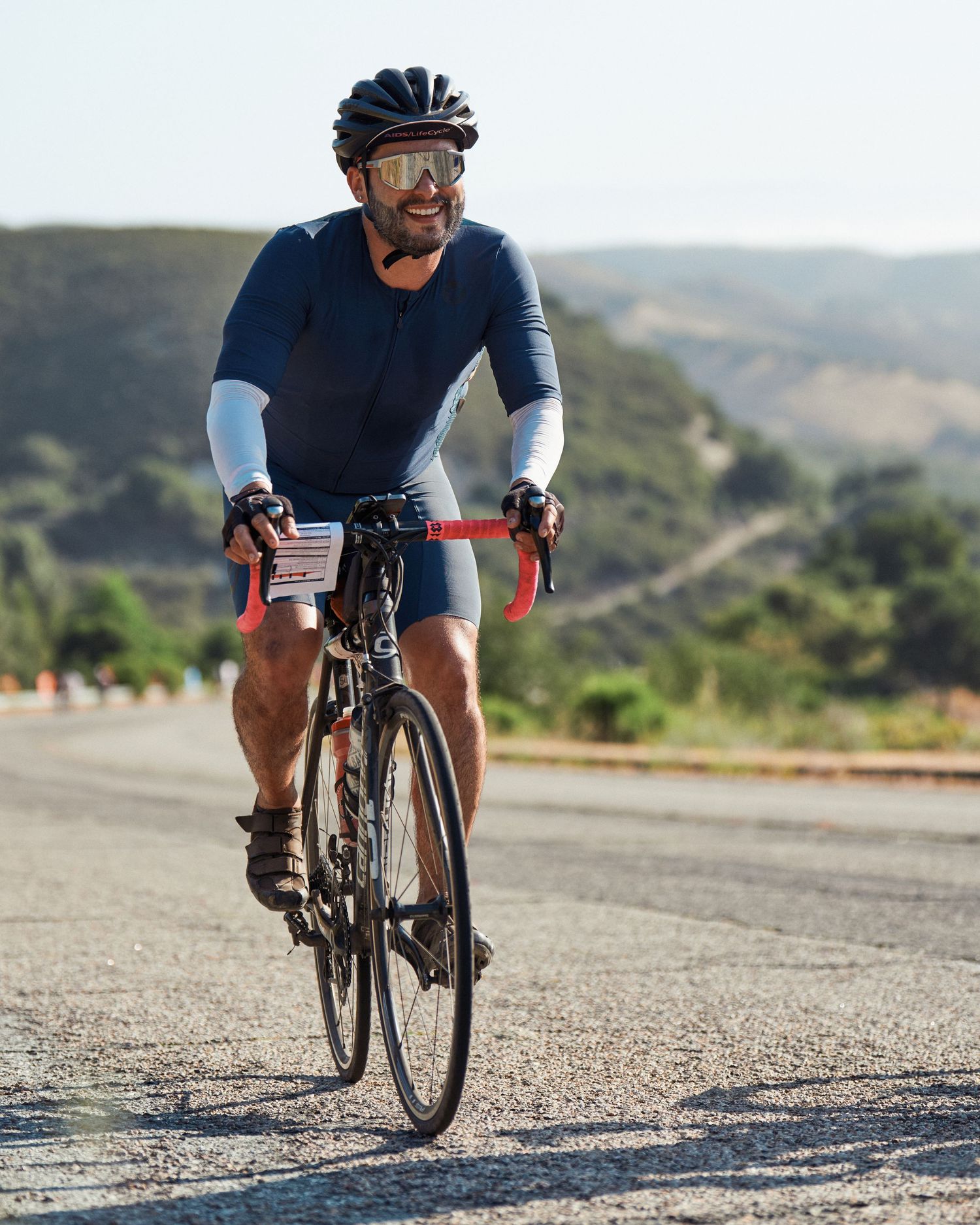 A cyclist in dark cycling gear rides a road bike along a scenic mountain road on a sunny day.