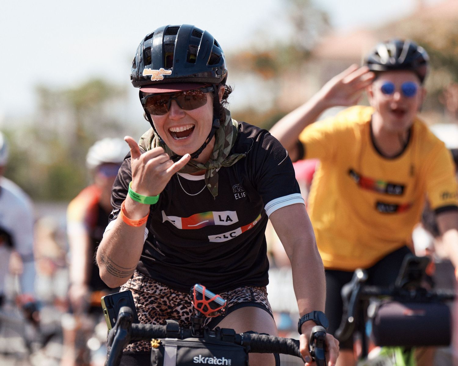 Cyclist in black helmet and sunglasses smiles while riding in a race with another rider visible in yellow behind.