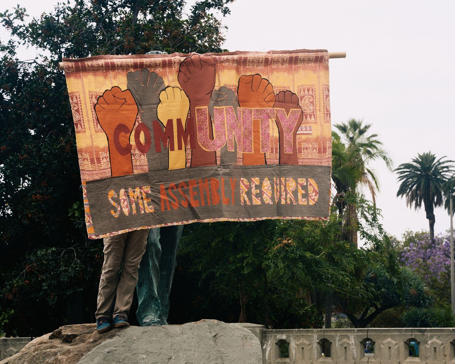 Demonstrator holds handmade “Community: Some Assembly Required” banner at MacArthur Park during May Day rally, Los Angeles, May 1, 2026.