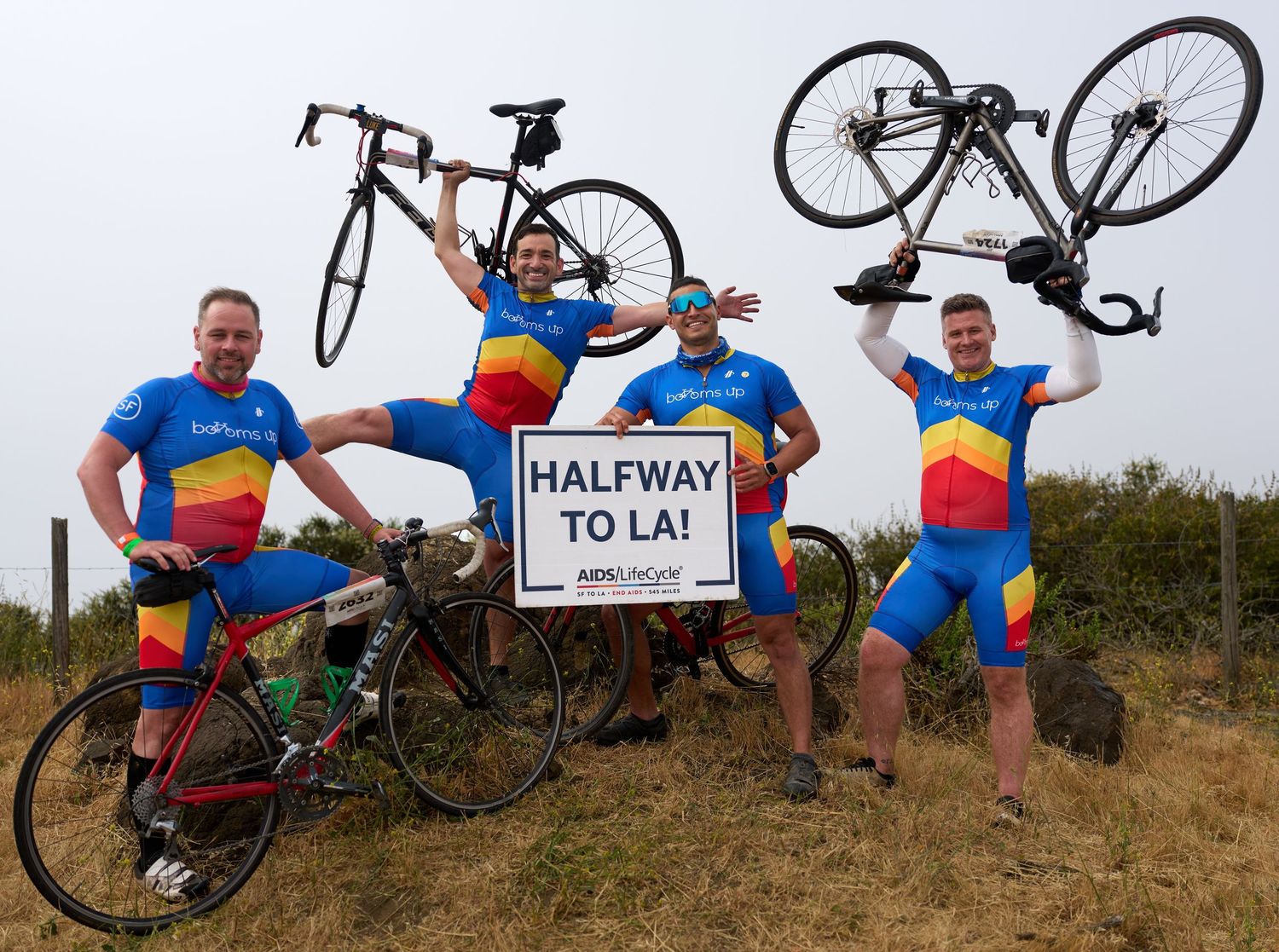 Cyclists in colorful Romanian flag jerseys celebrate with bikes and a 'Halfway to LA' sign on rocky terrain.