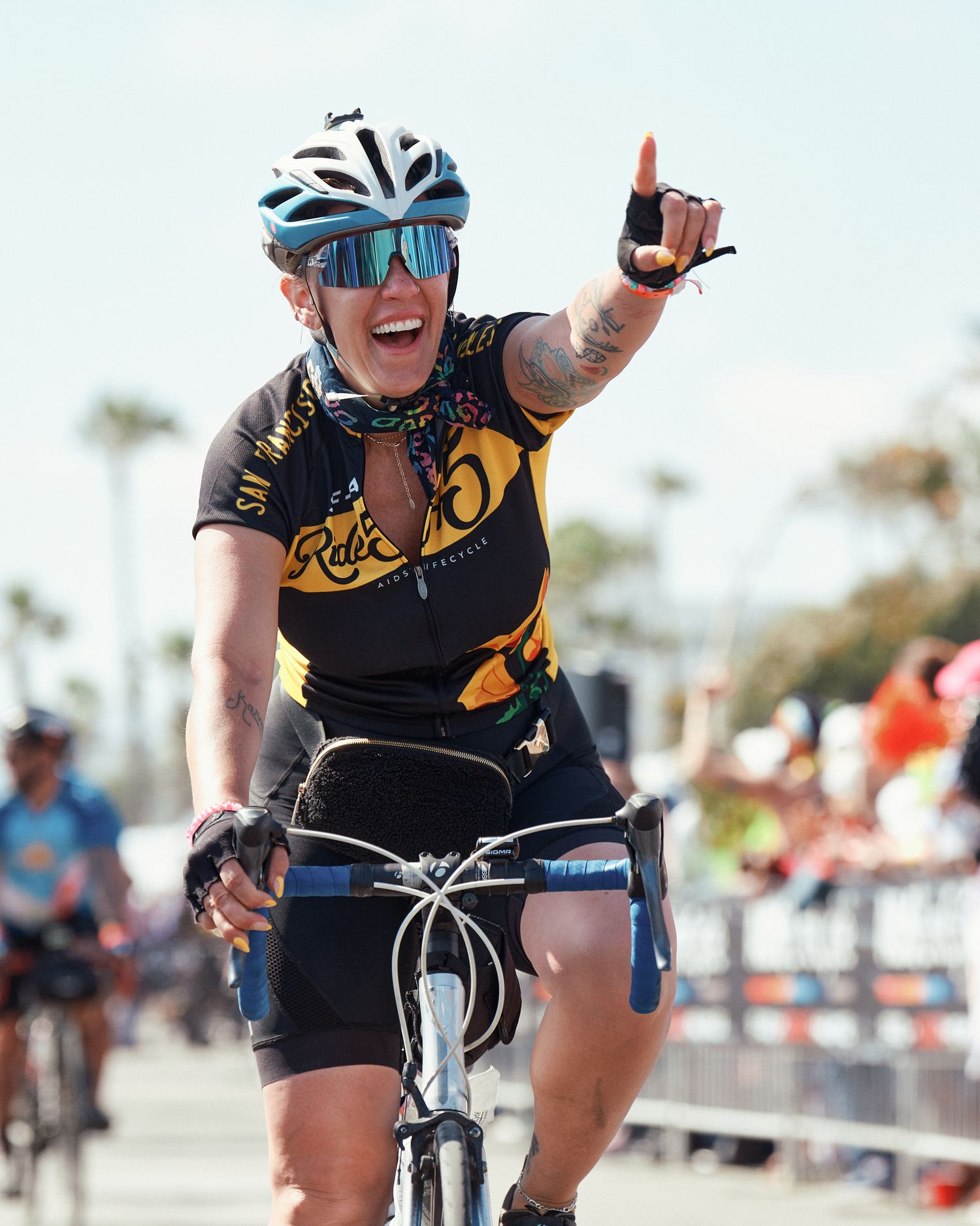 Cyclist wearing blue helmet and yellow-black cycling jersey celebrates with thumbs up gesture while riding bicycle.
