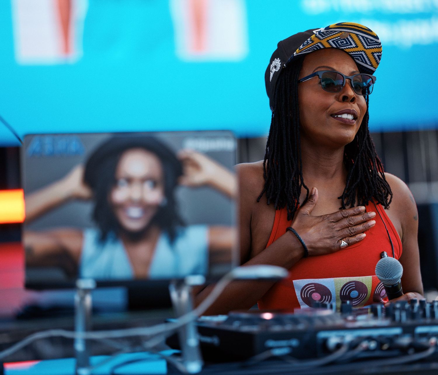 DJ performs at an outdoor music festival wearing sunglasses and a cap while mixing on turntables in bright sunlight.