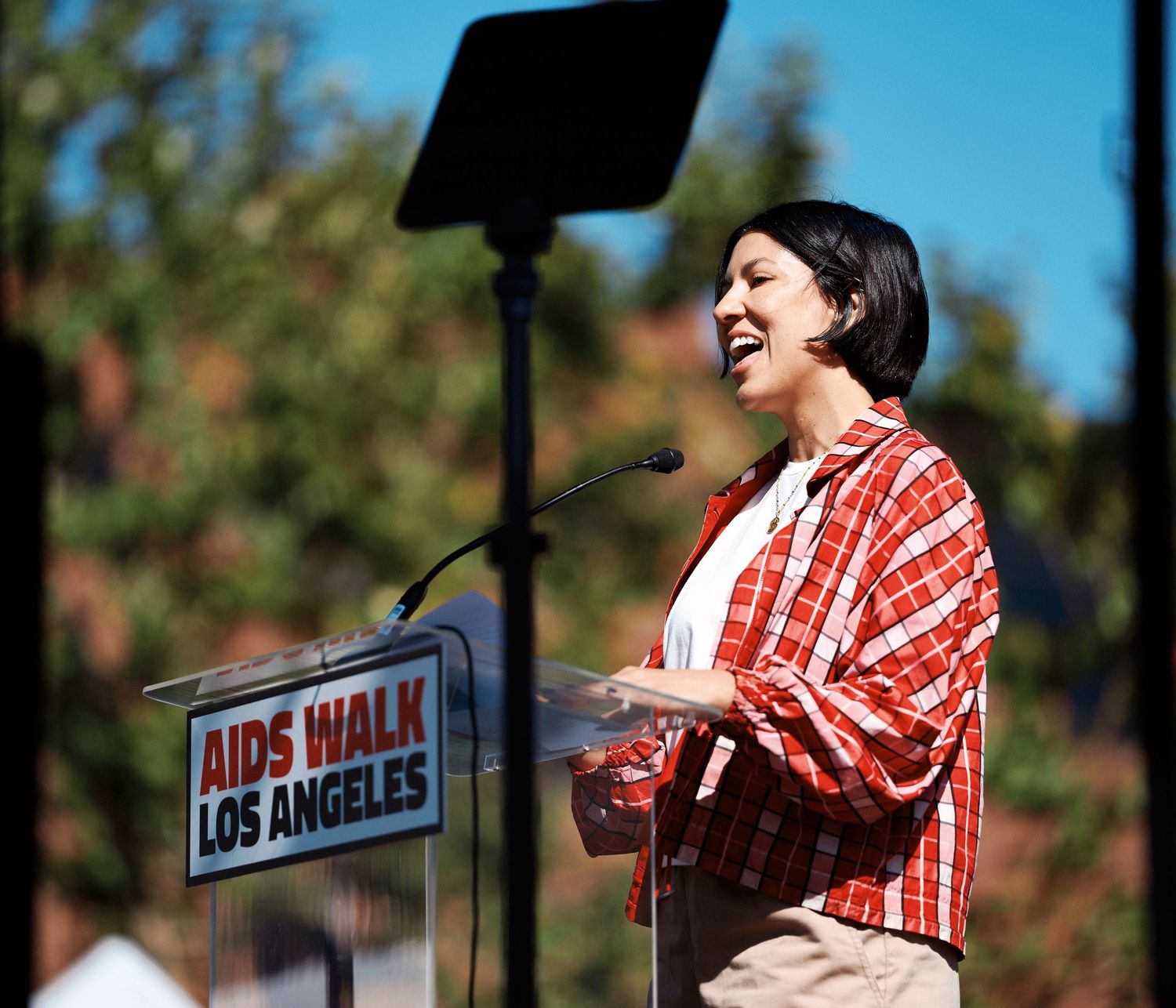 A performer in a red plaid shirt sings passionately on stage at an outdoor event in Los Angeles.