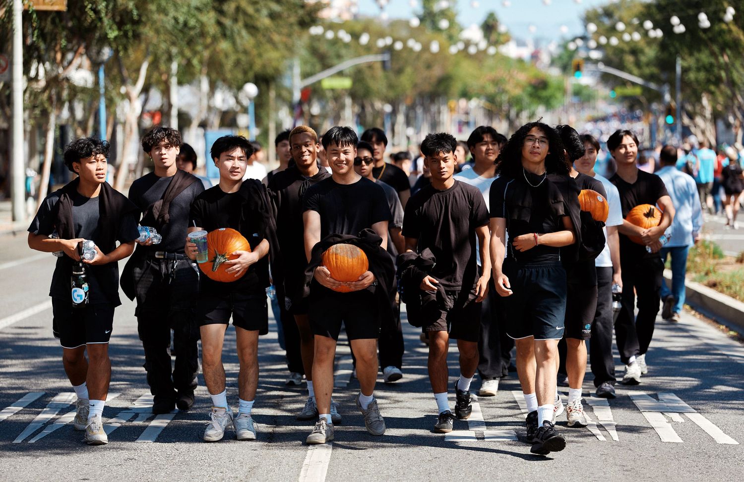 Large group of people wearing black clothing sit together on a sunny sidewalk during an outdoor gathering.