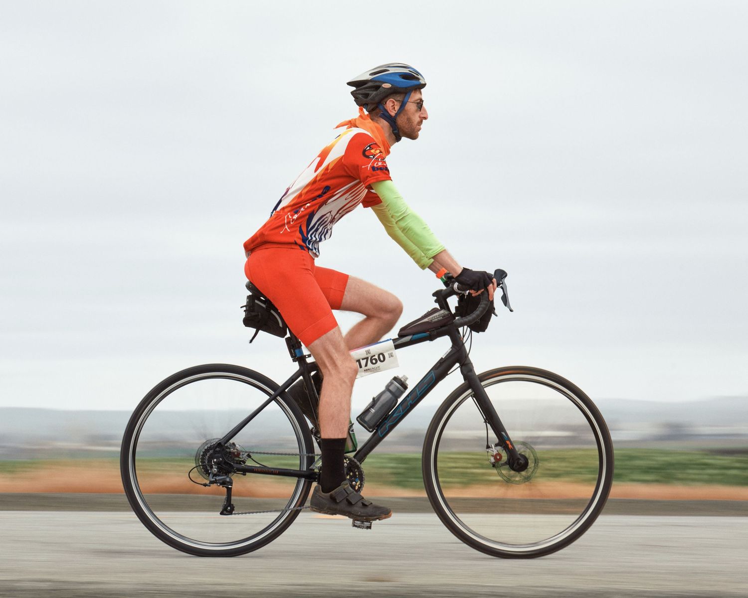 Cyclist in orange and yellow jersey rides road bike through scenic countryside landscape.