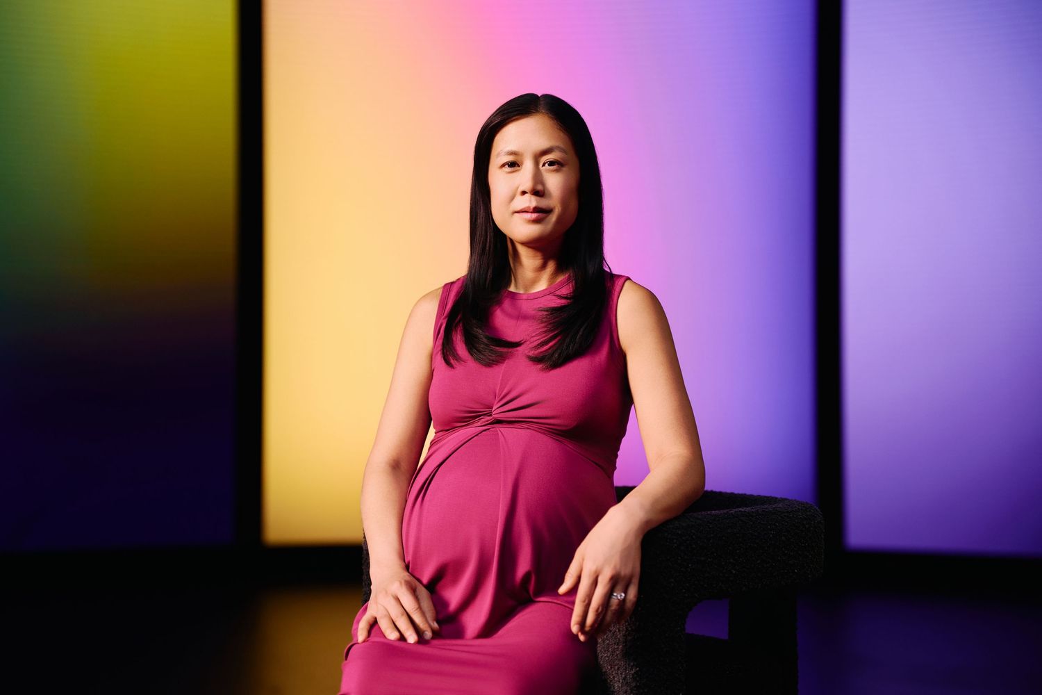 A pregnant woman in a magenta dress sits against a colorful rainbow gradient background for a portrait session.
