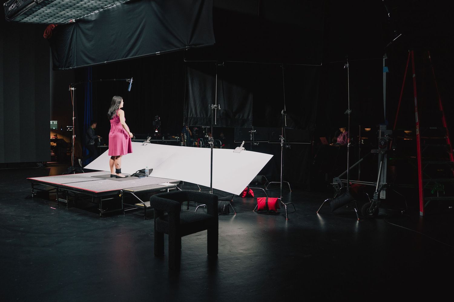 Someone in a pink dress stands alone on a brightly lit stage platform in a dark theater setting.