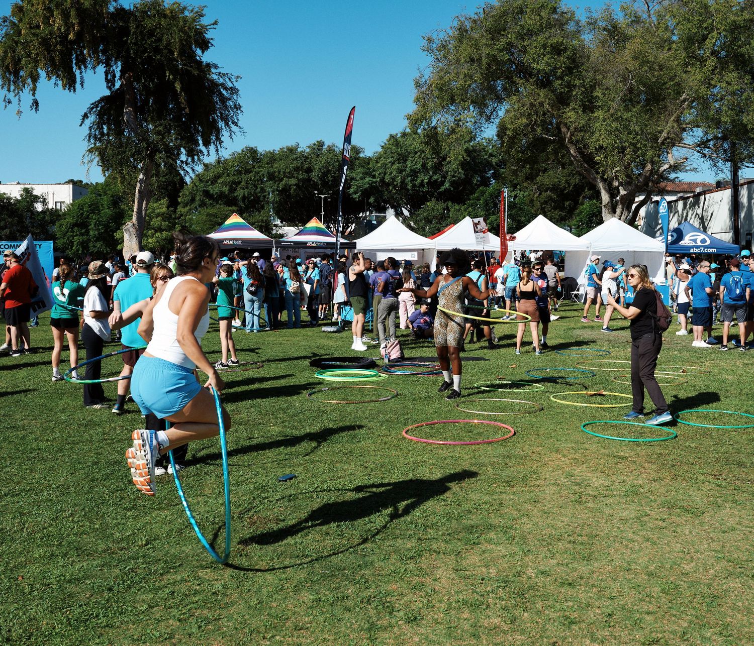 People hula hooping on a grassy field at an outdoor festival with white tents and trees in the background.