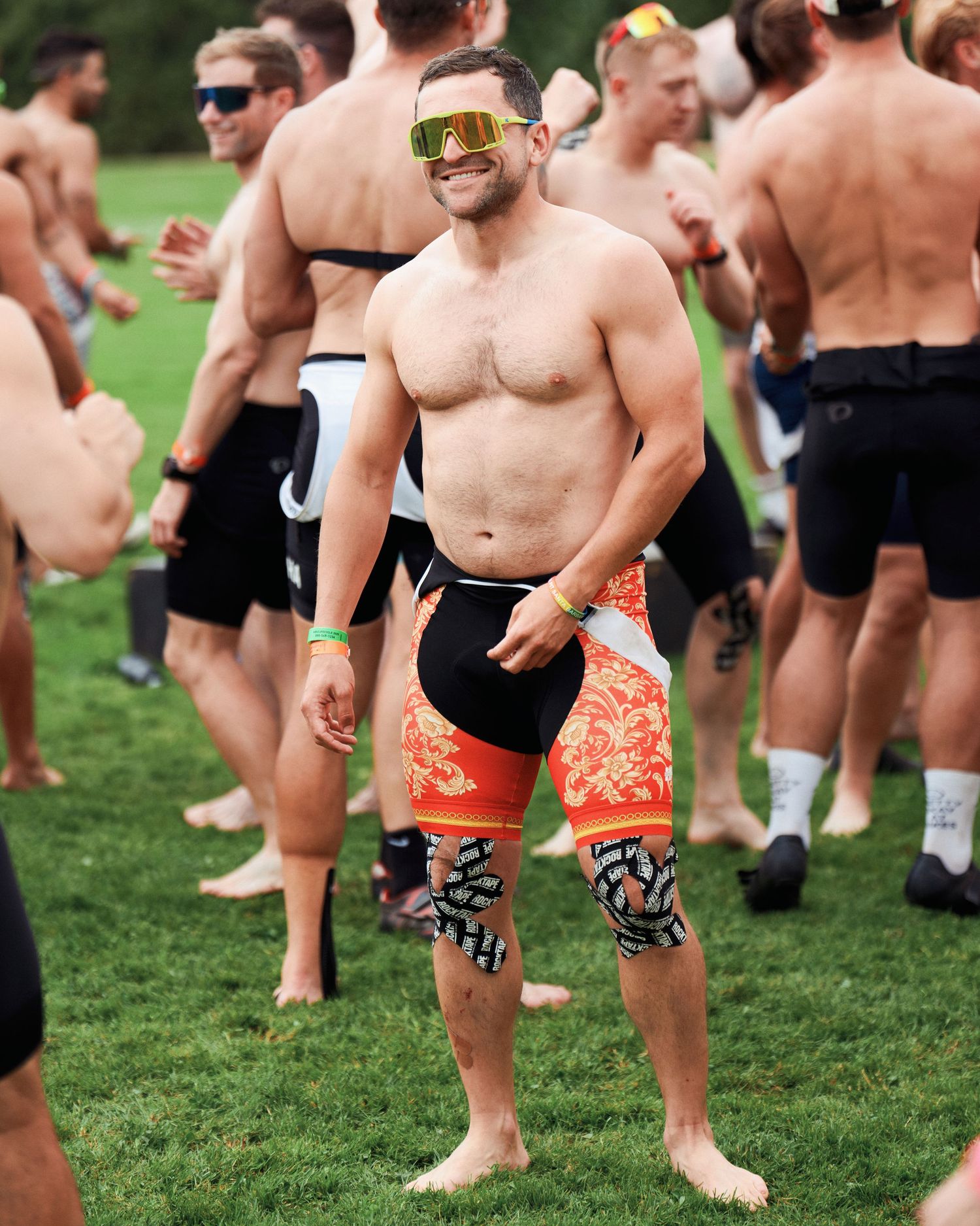 Shirtless athlete wearing sports sunglasses and patterned shorts at outdoor event.