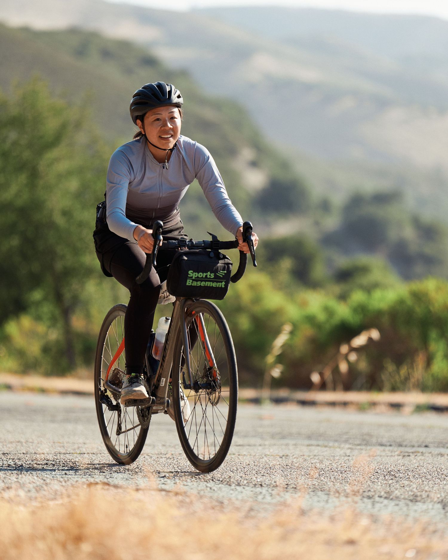 Cyclist in gray athletic wear biking along a scenic mountain road with handlebar bag on a gravel bike.
