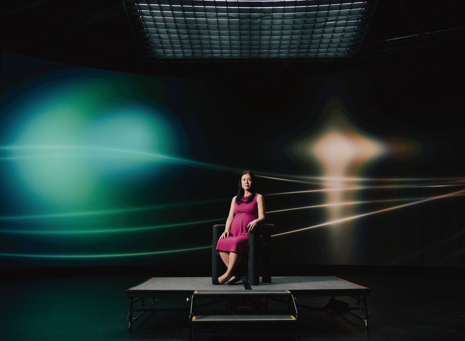 Figure in pink dress sits on platform against dramatic backdrop with teal lighting and golden light beams.