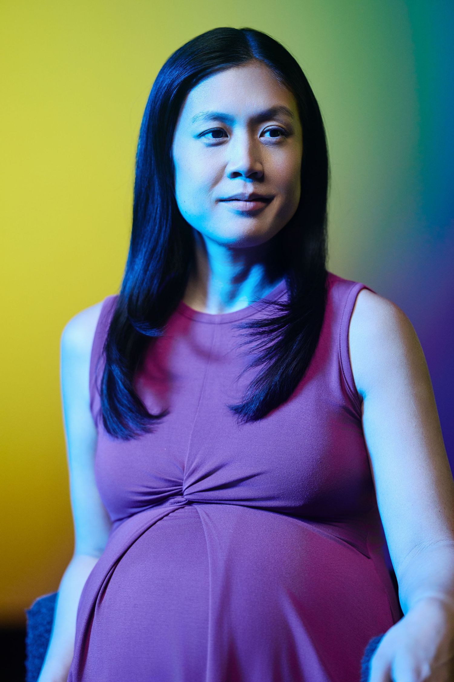 A pregnant person in a purple sleeveless top poses against a colorful gradient background of yellow and teal.