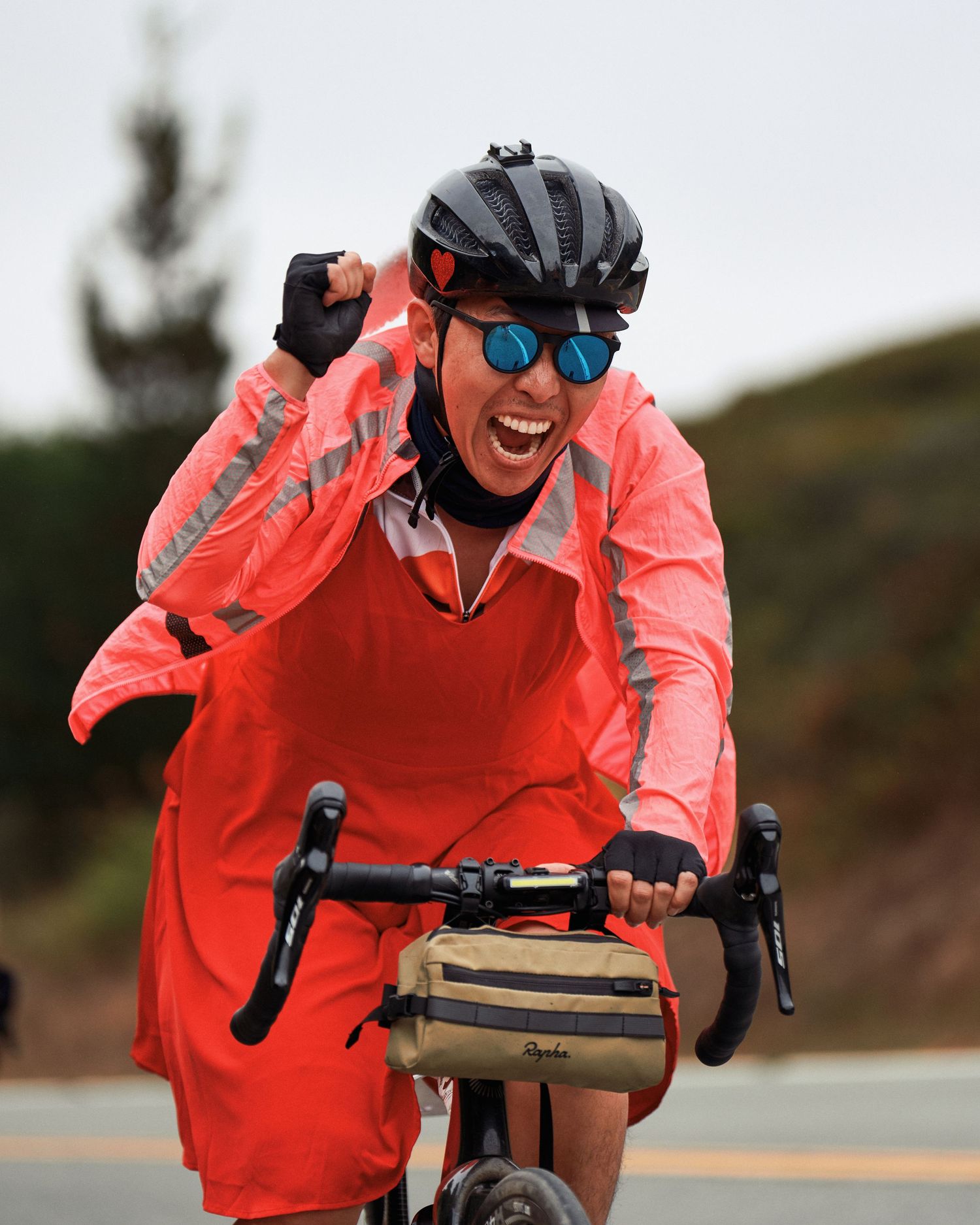 Cyclist in bright coral-colored jacket smiling while riding bike on coastal road.