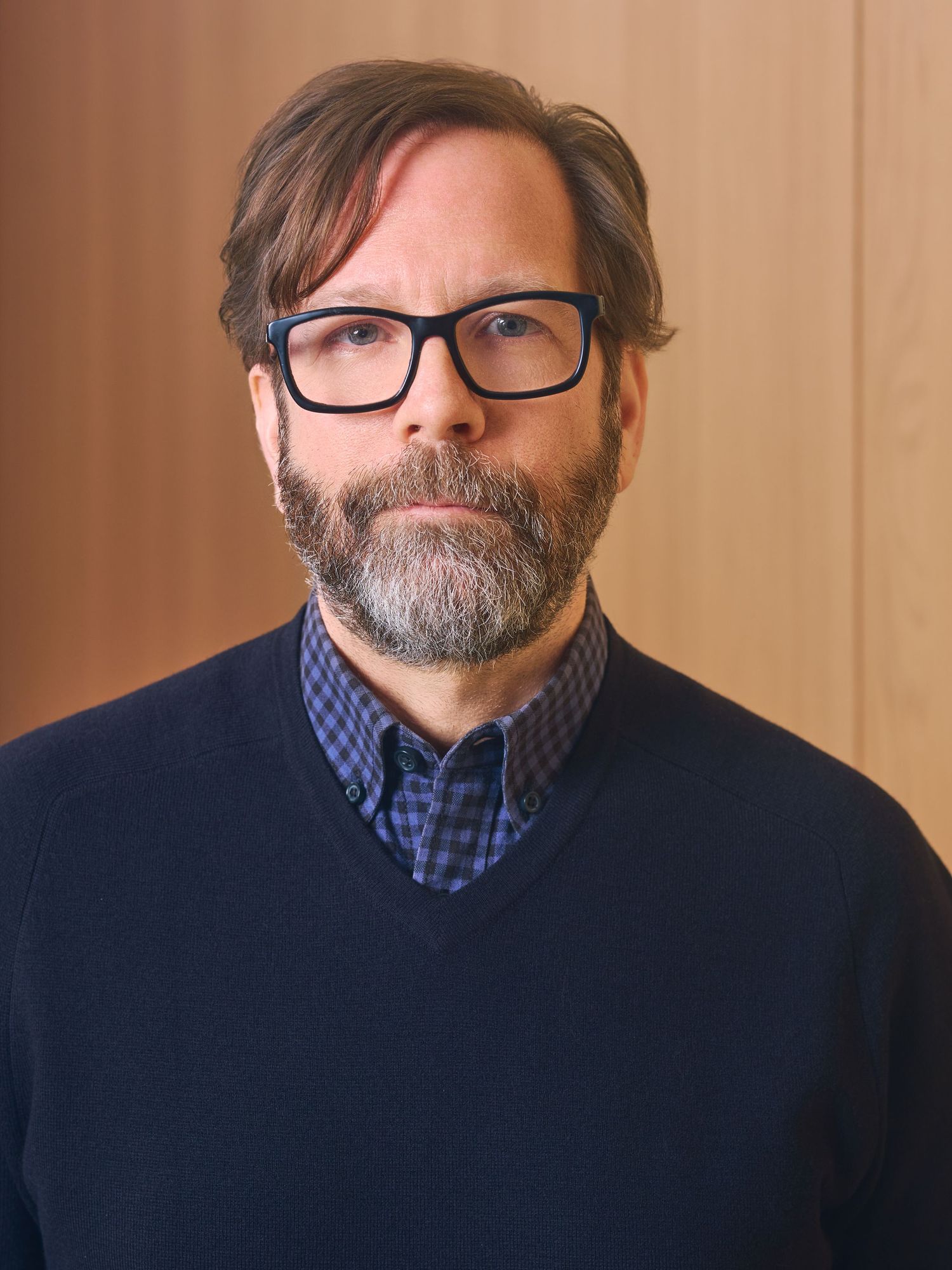 Middle-aged man with glasses and gray beard wearing navy sweater over plaid shirt, posed against wooden background.