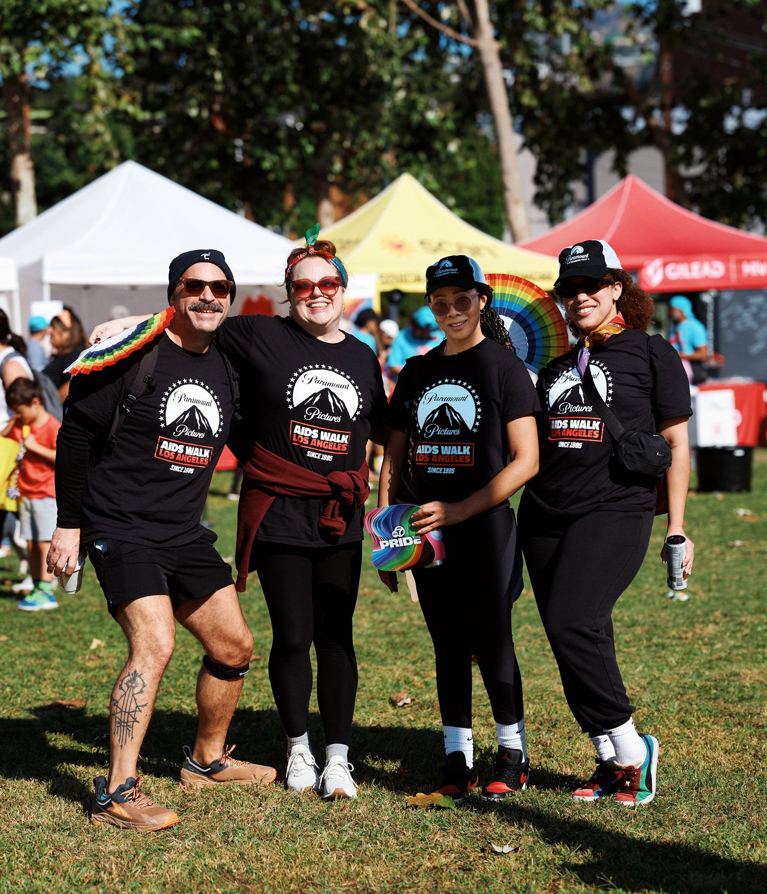 A line of people wearing matching black t-shirts with logos kneel and crouch on grass at an outdoor event.