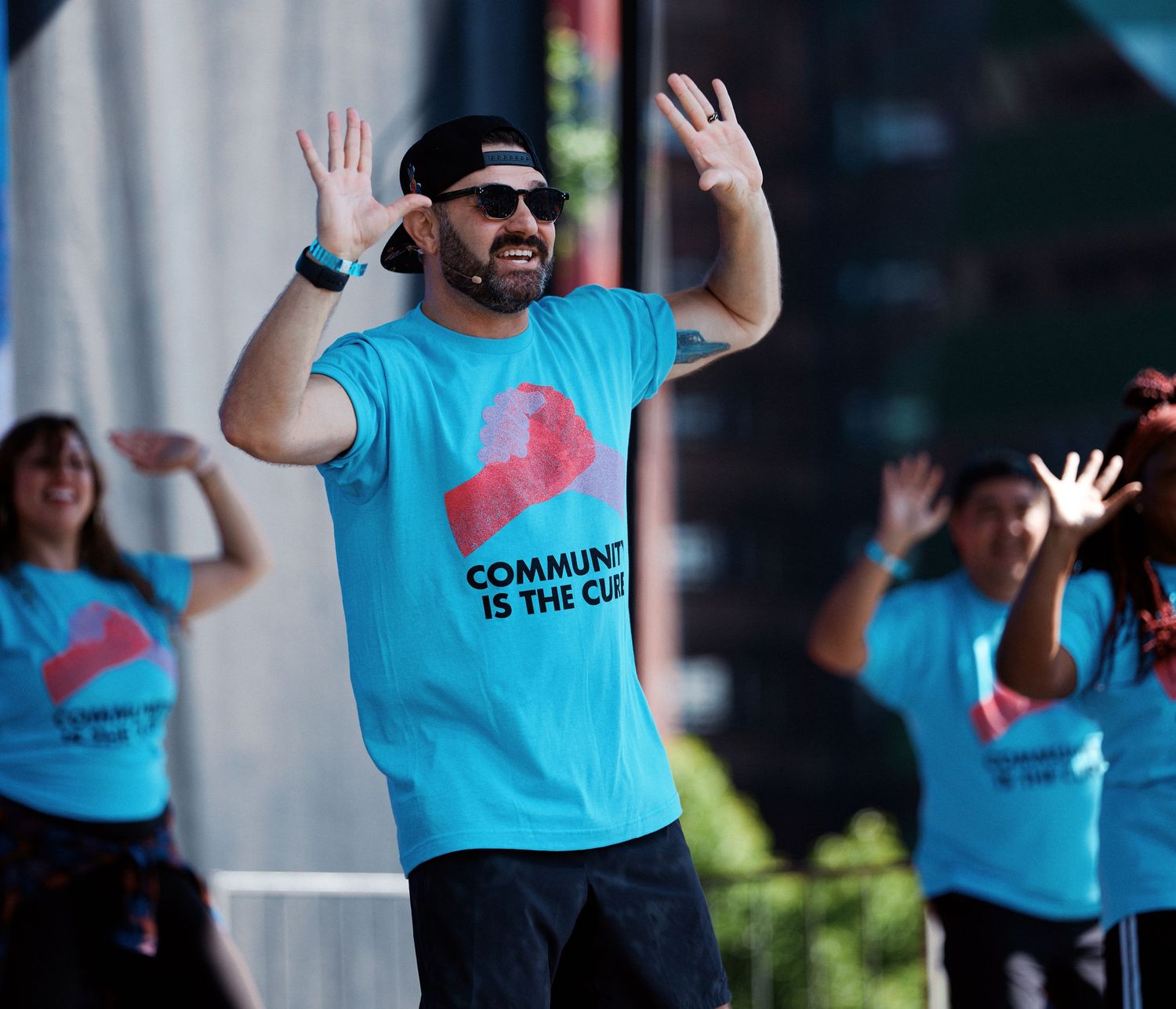A group of people in matching light blue t-shirts wave and smile while participating in an outdoor event.