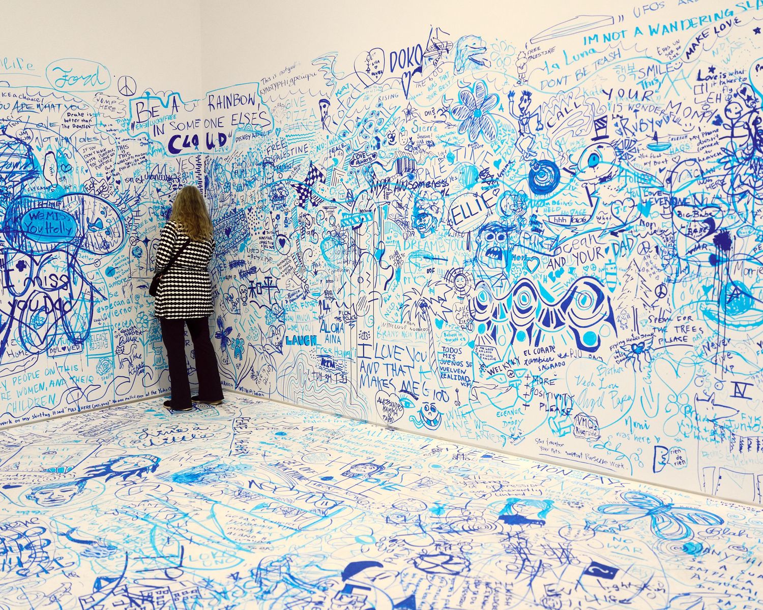 Woman standing in a room covered floor-to-ceiling with blue marker graffiti art and text in a gallery installation.