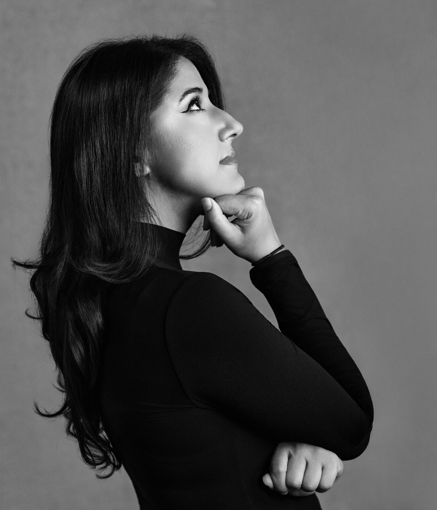 Black and white portrait of a thoughtful young woman in turtleneck, hand on chin, looking upward.