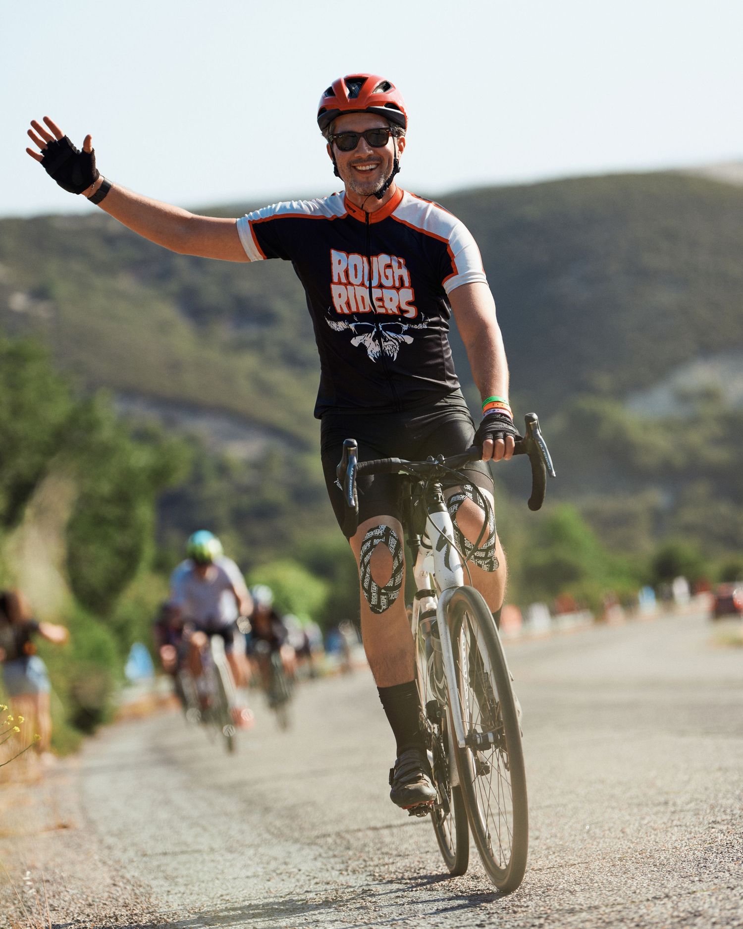 Cyclist in black t-shirt waving while riding on a mountain road with rolling hills in the background.