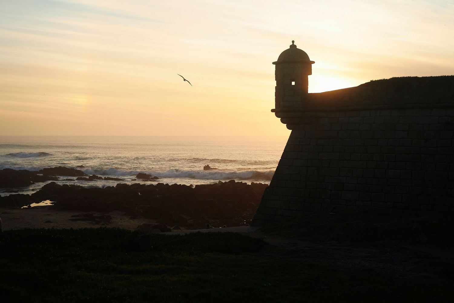 Panoramic sunset view over a rocky coastline with waves crashing against the shore and silhouetted cliffs in the distance.