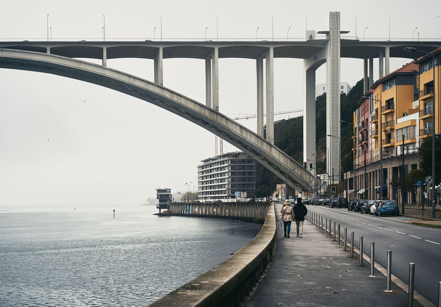 Curved concrete arch bridge spans over waterfront promenade with modern buildings and coastline in Porto, Portugal.