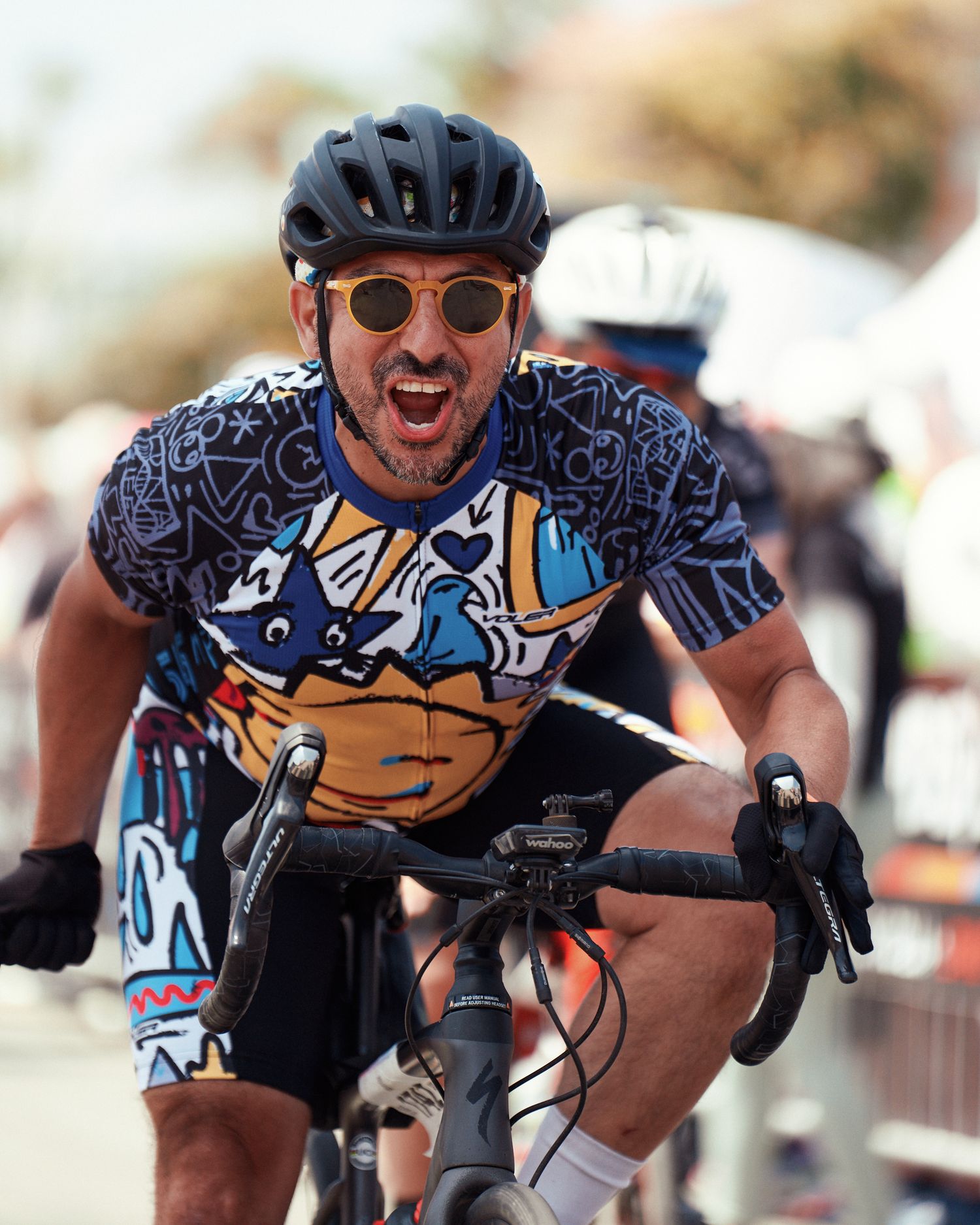 Cyclist in a patterned blue jersey and black helmet shows excitement while riding during a competition.