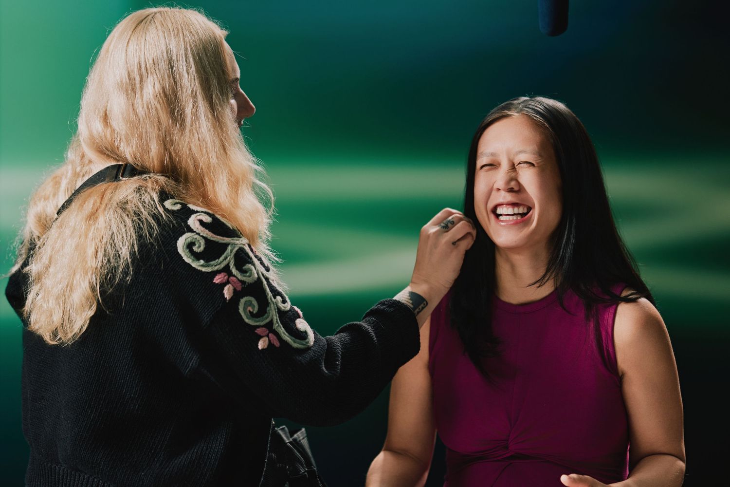 A makeup artist applies cosmetics to a laughing person wearing a burgundy sleeveless top against a green backdrop.