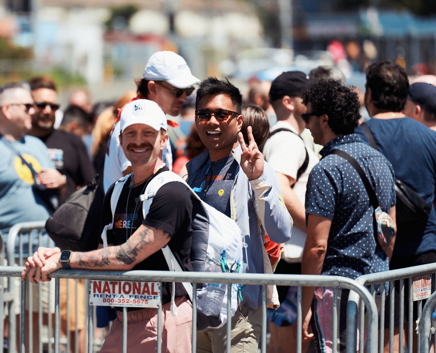 A diverse crowd of people gathered behind safety barriers at an outdoor event, with some wearing sunglasses and hats.