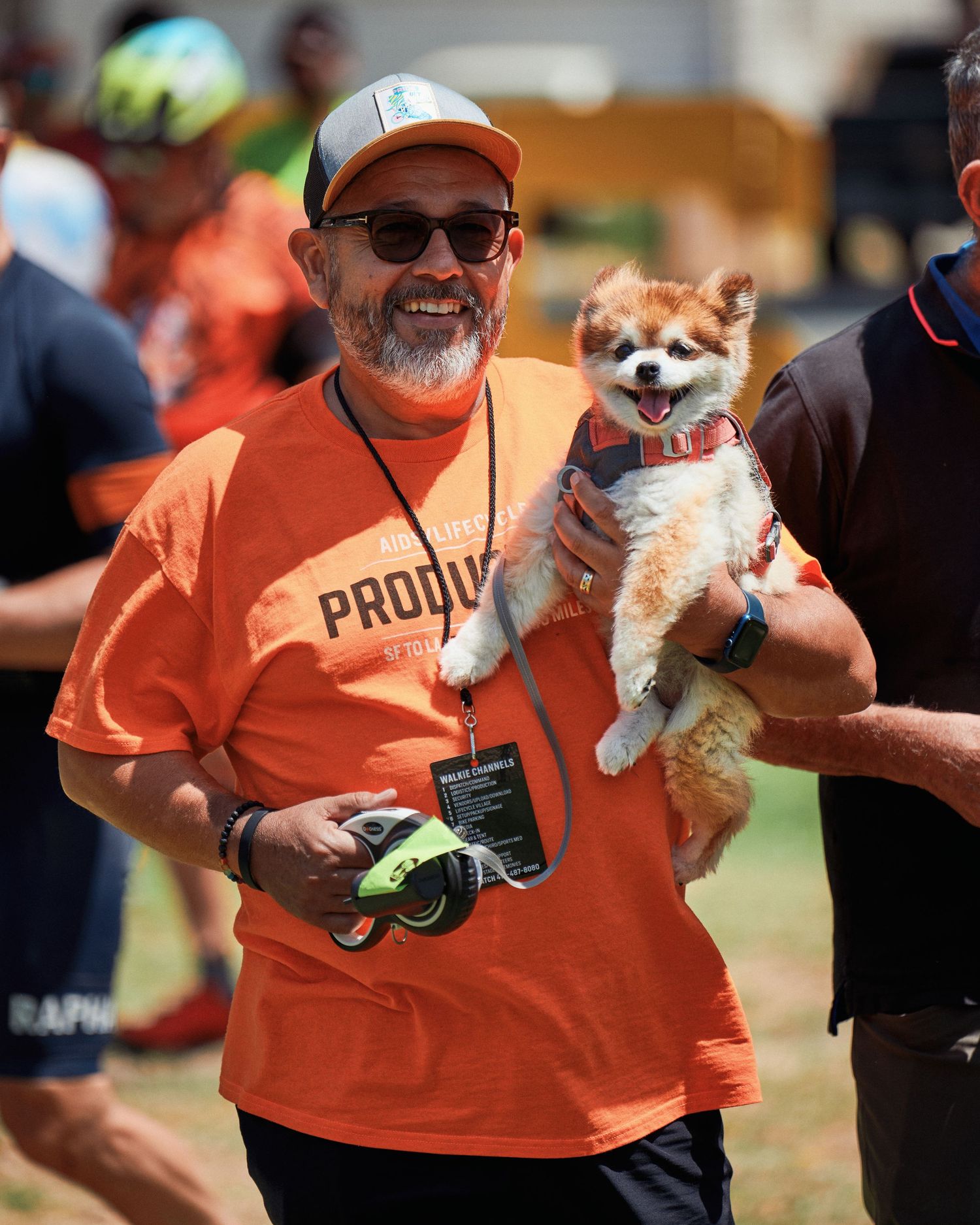 Someone in orange t-shirt smiling while holding a happy Pomeranian dog at outdoor event.