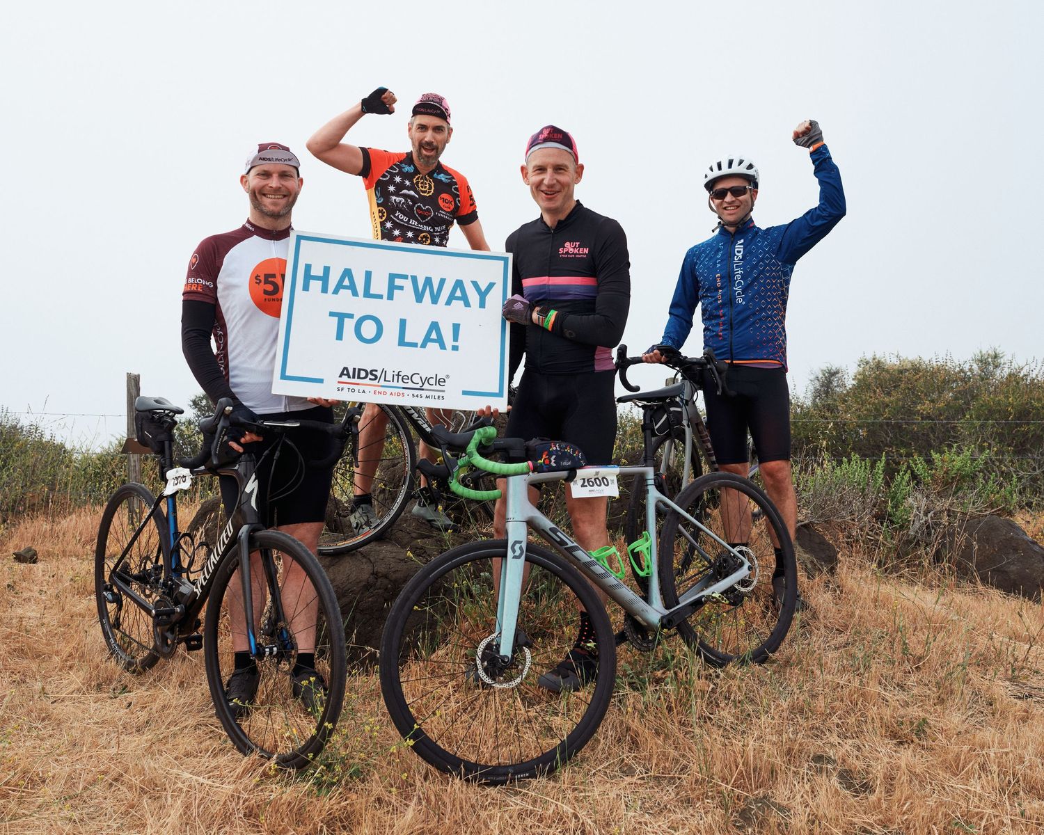 Cyclists posing with 'Halfway to LA' sign on a dirt trail with their bikes.