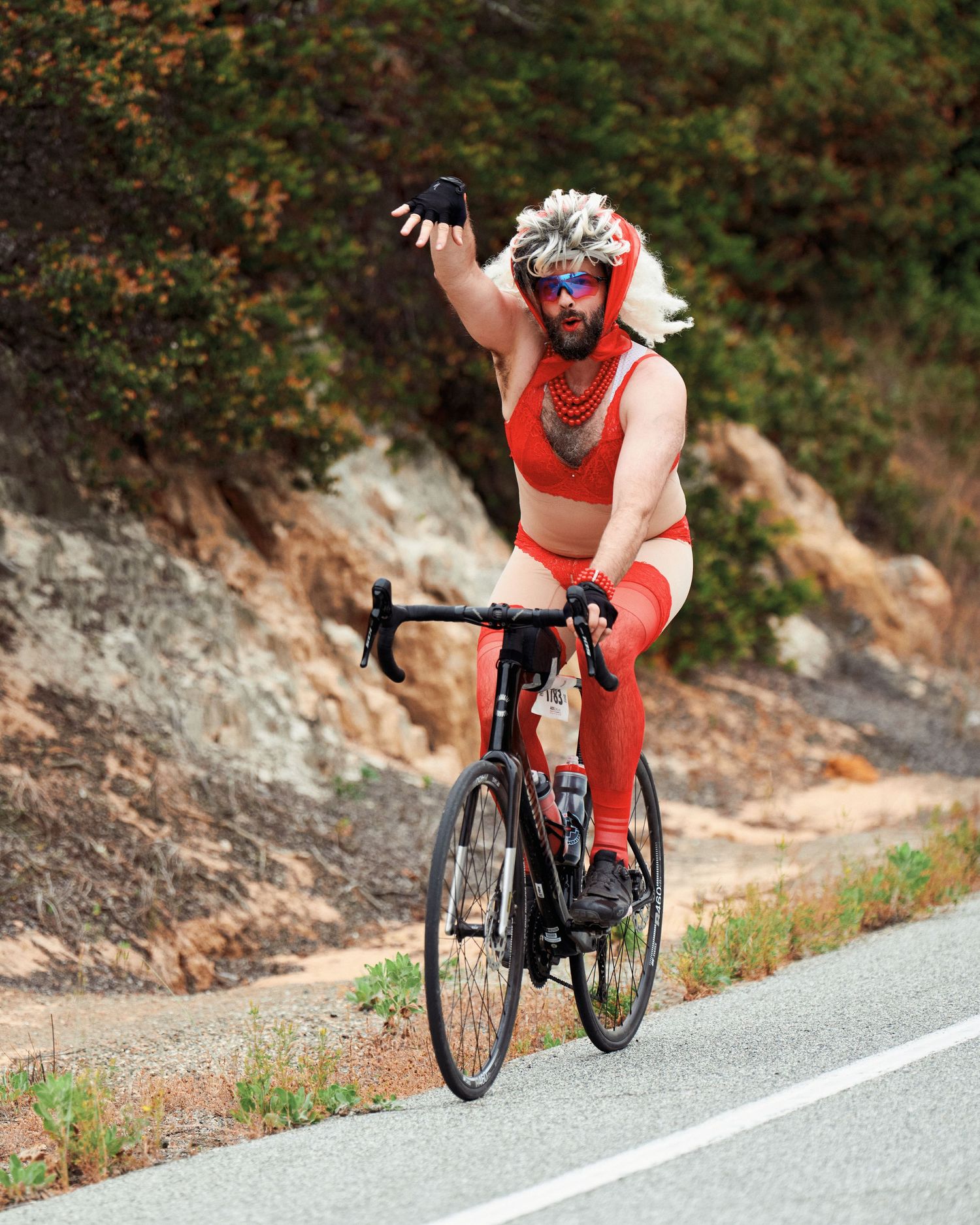 Cyclist in red outfit performs celebratory gesture while riding bike on mountain road.