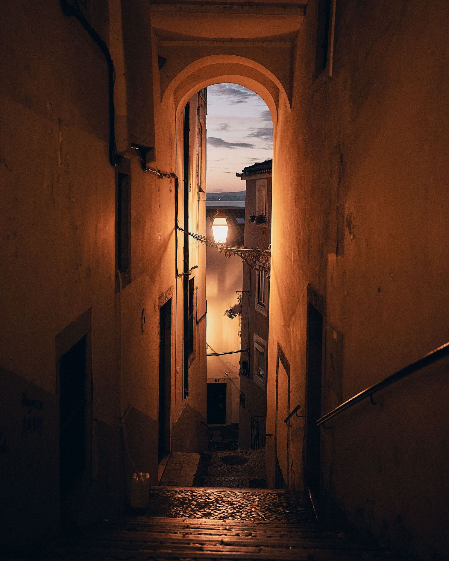 Narrow cobblestone alley with arched passageway and warm lantern light illuminating old European buildings at dusk.