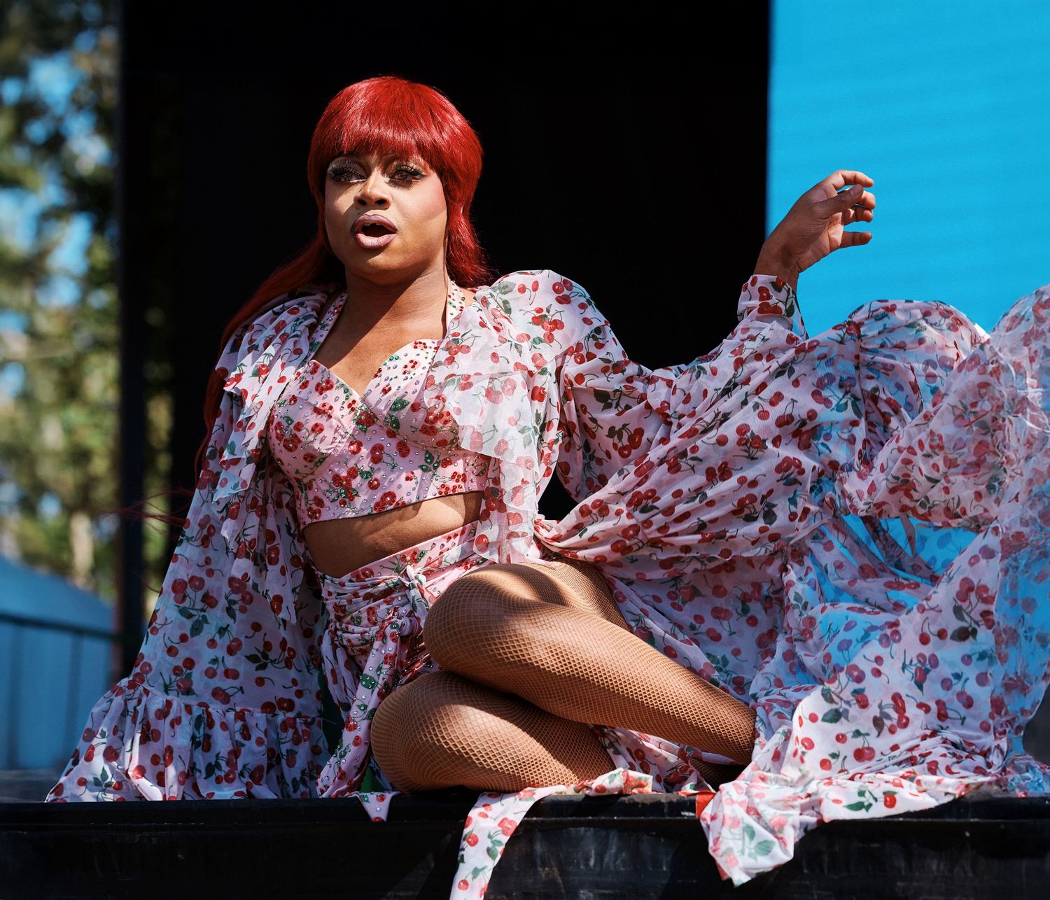 A person in a floral dress with flowing sleeves poses against a bright blue sky and trees.