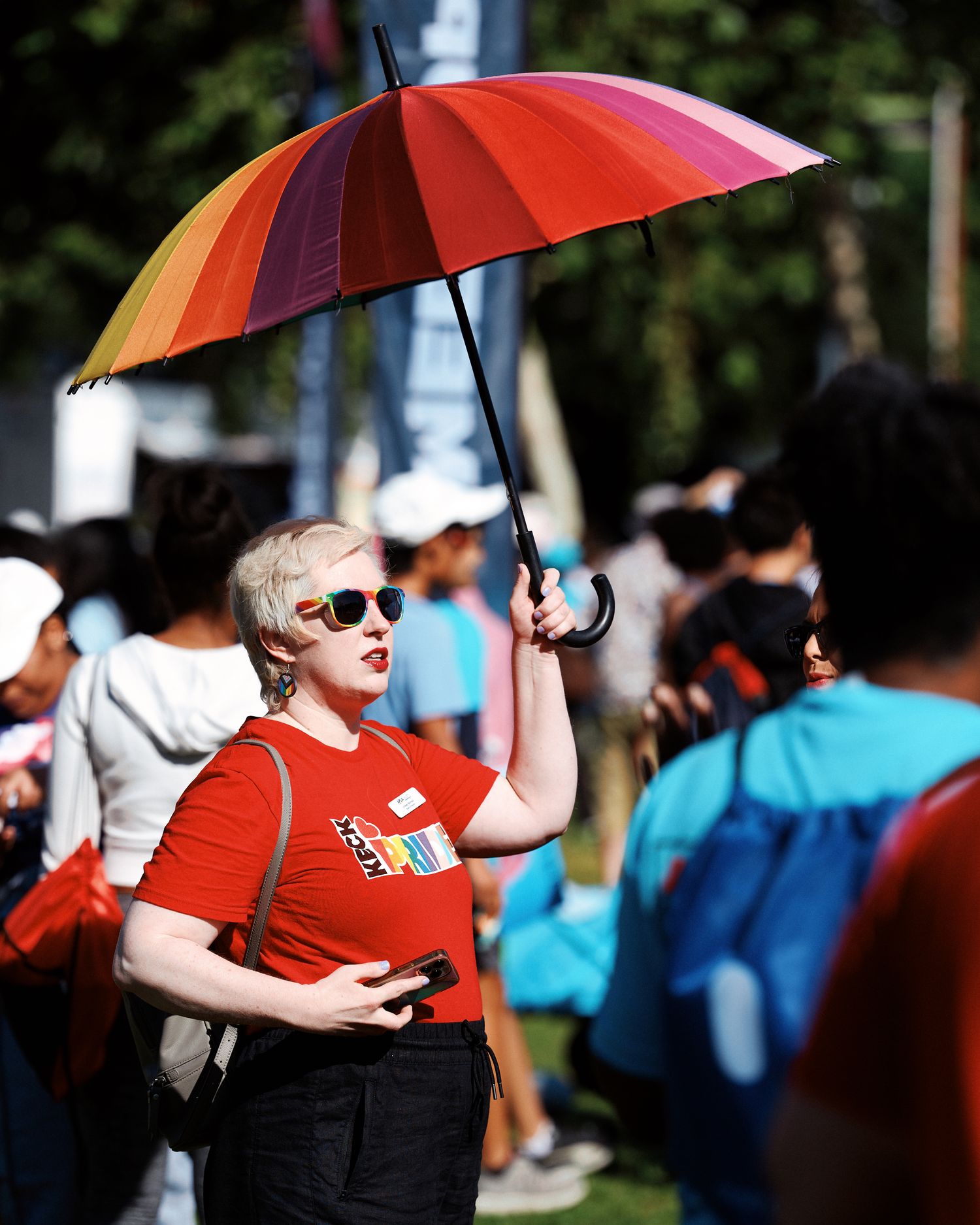 A person in red holds a colorful rainbow umbrella during a sunny outdoor event with a crowd in the background.